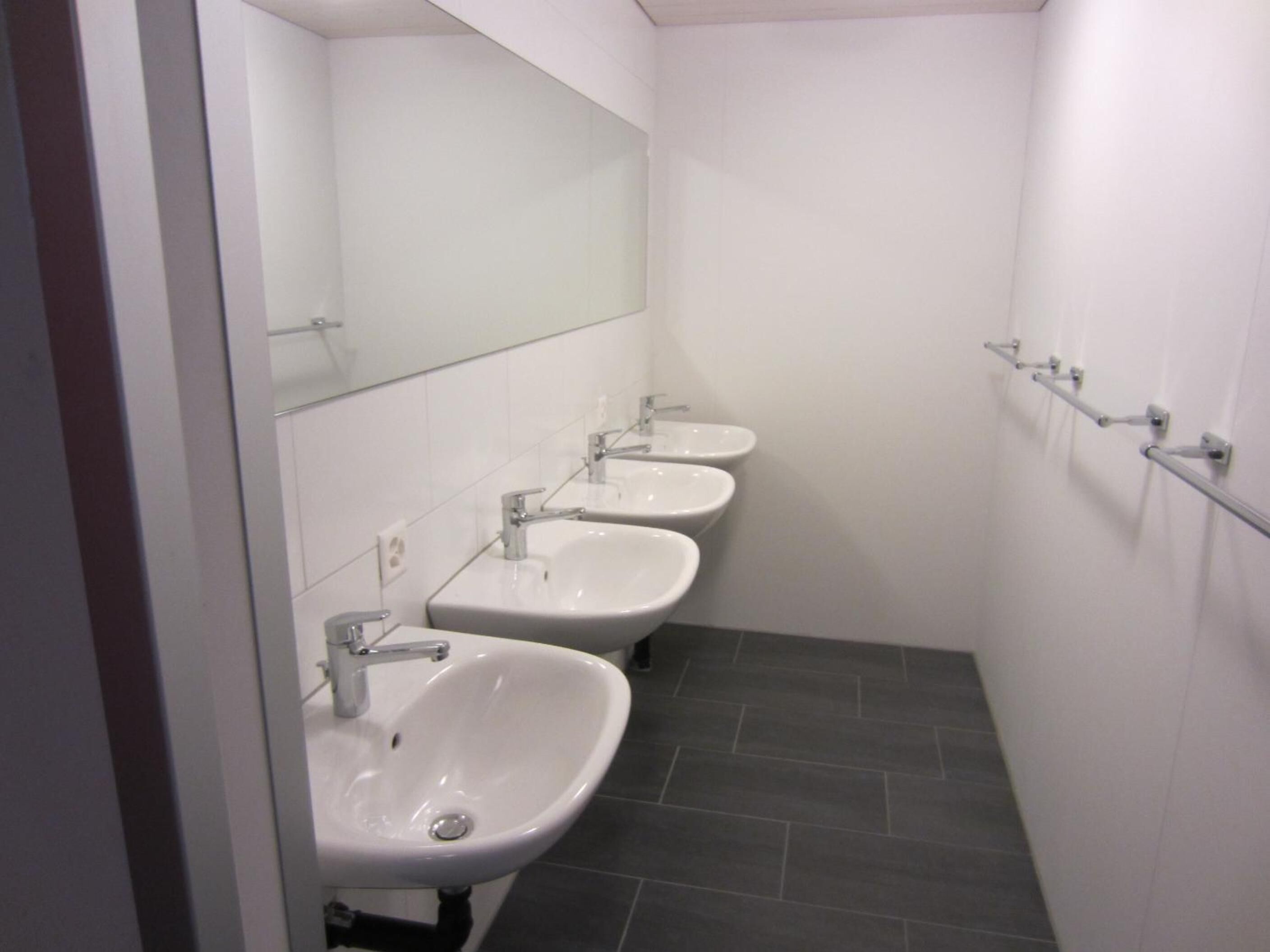 A narrow bathroom with three white sinks and a mirror above them.