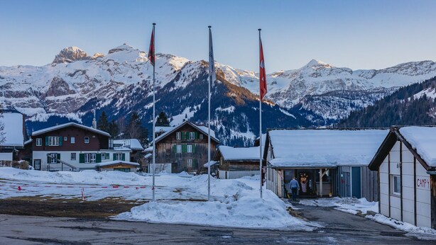 Un pueblo cubierto de nieve con casas de madera tradicionales, un pequeño edificio con el rótulo "CAMP 4" y banderas en los mástiles.