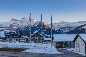A snow-covered mountain village with traditional wooden houses, a small building labeled "CAMP 4", and flags on flagpoles. - Mountain Lodge Backpackercamp (Lenk)