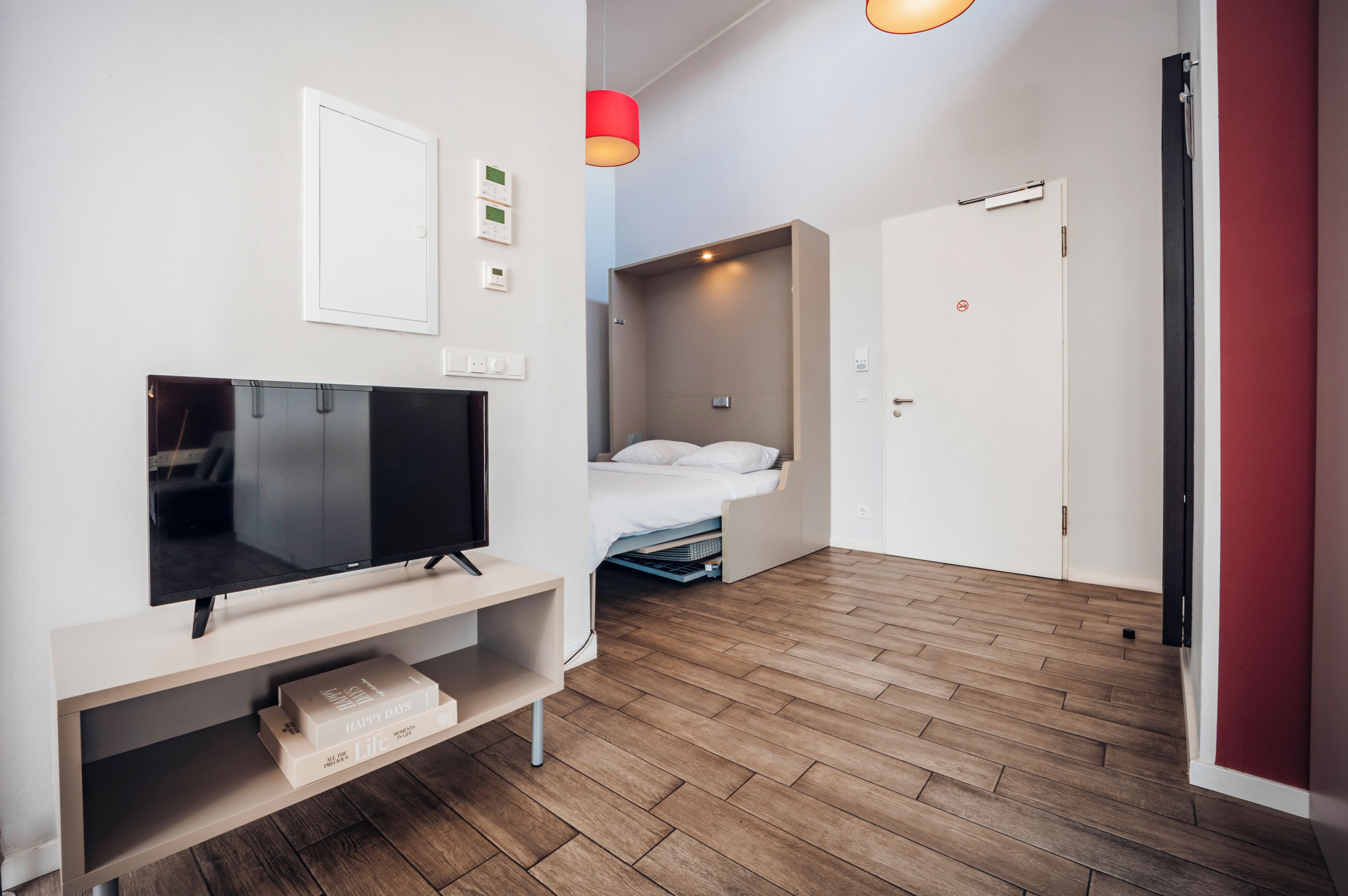A modern hotel room with a flat-screen TV, a bed, a built-in wardrobe, and a door leading to another room.