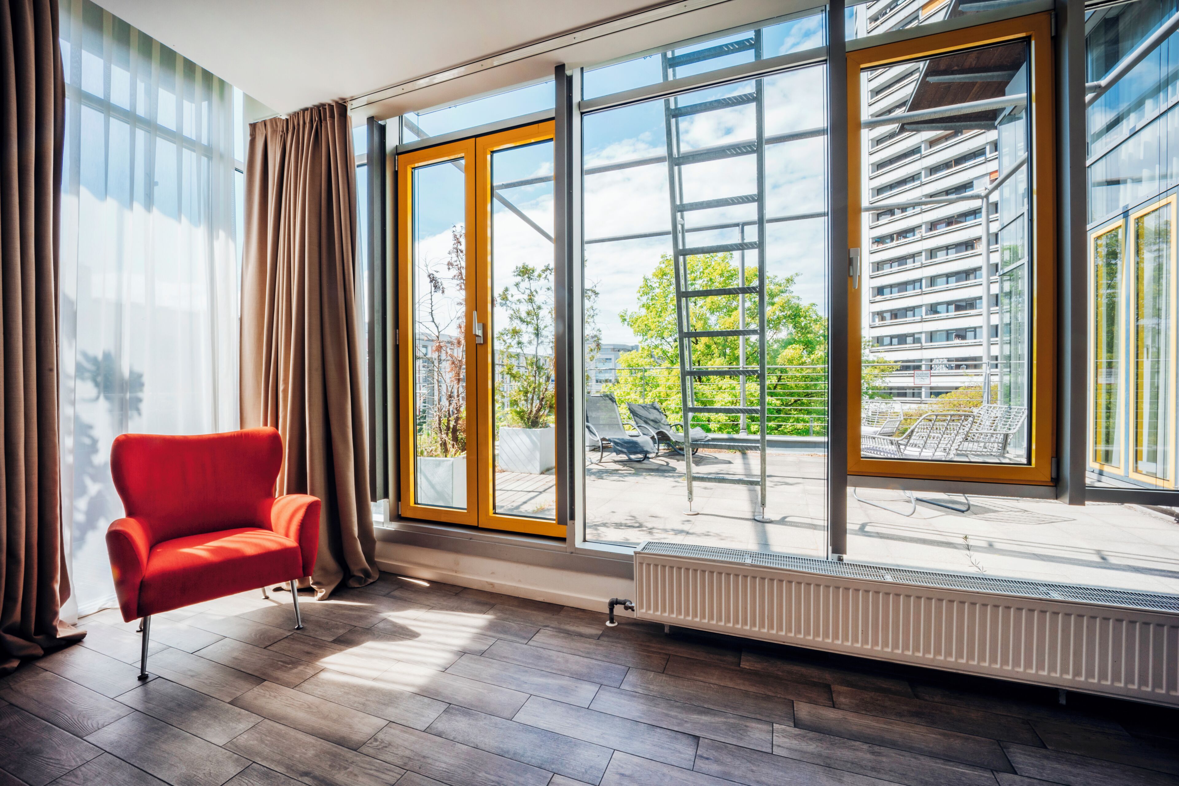 Ein modernes Zimmer mit einem roten Sessel, großen Fenstern und Blick auf ein Gebäude und Grünflächen.