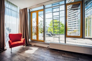 A modern room with a red chair, large windows, and a view of a building and greenery outside. - Brera Serviced Apartments Munich West (Munich)