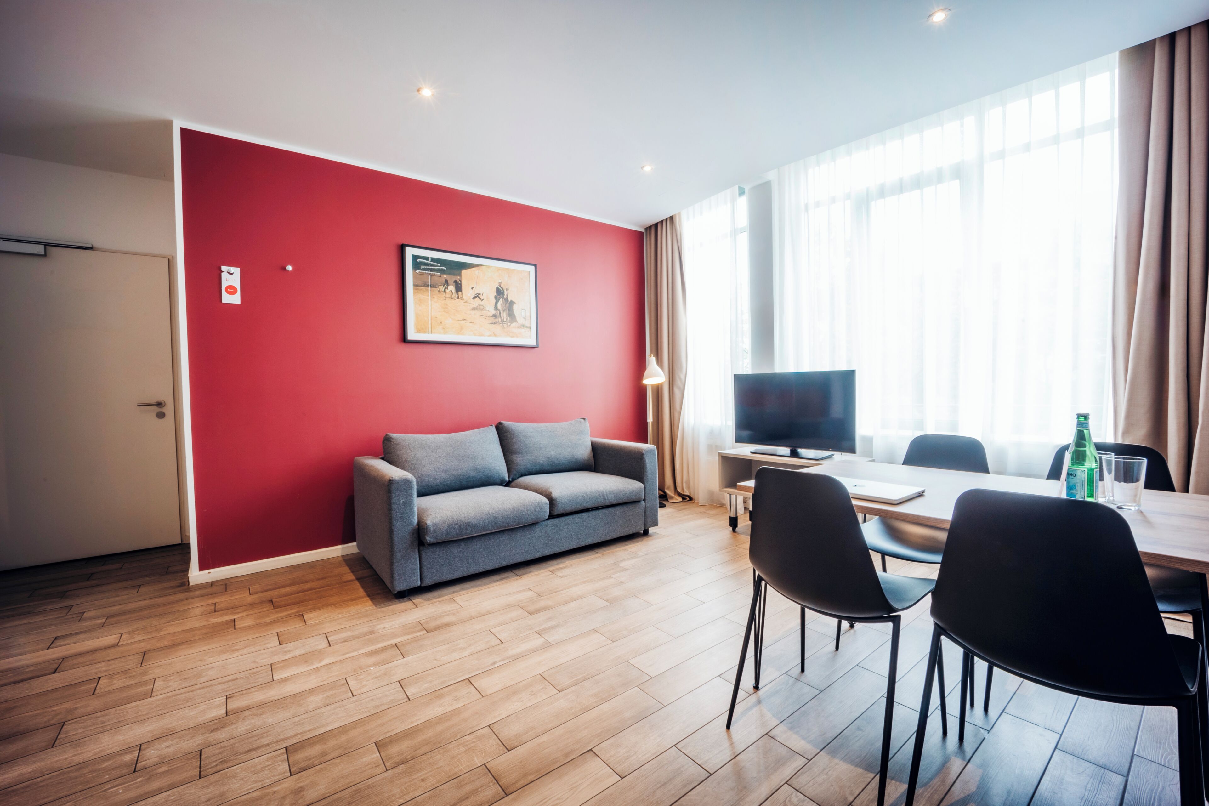 A modern living room with a red accent wall, a grey sofa, a flat-screen TV, and a dining area with a table and chairs.
