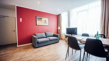 A modern living room with a red accent wall, a grey sofa, a flat-screen TV, and a dining area with a table and chairs.