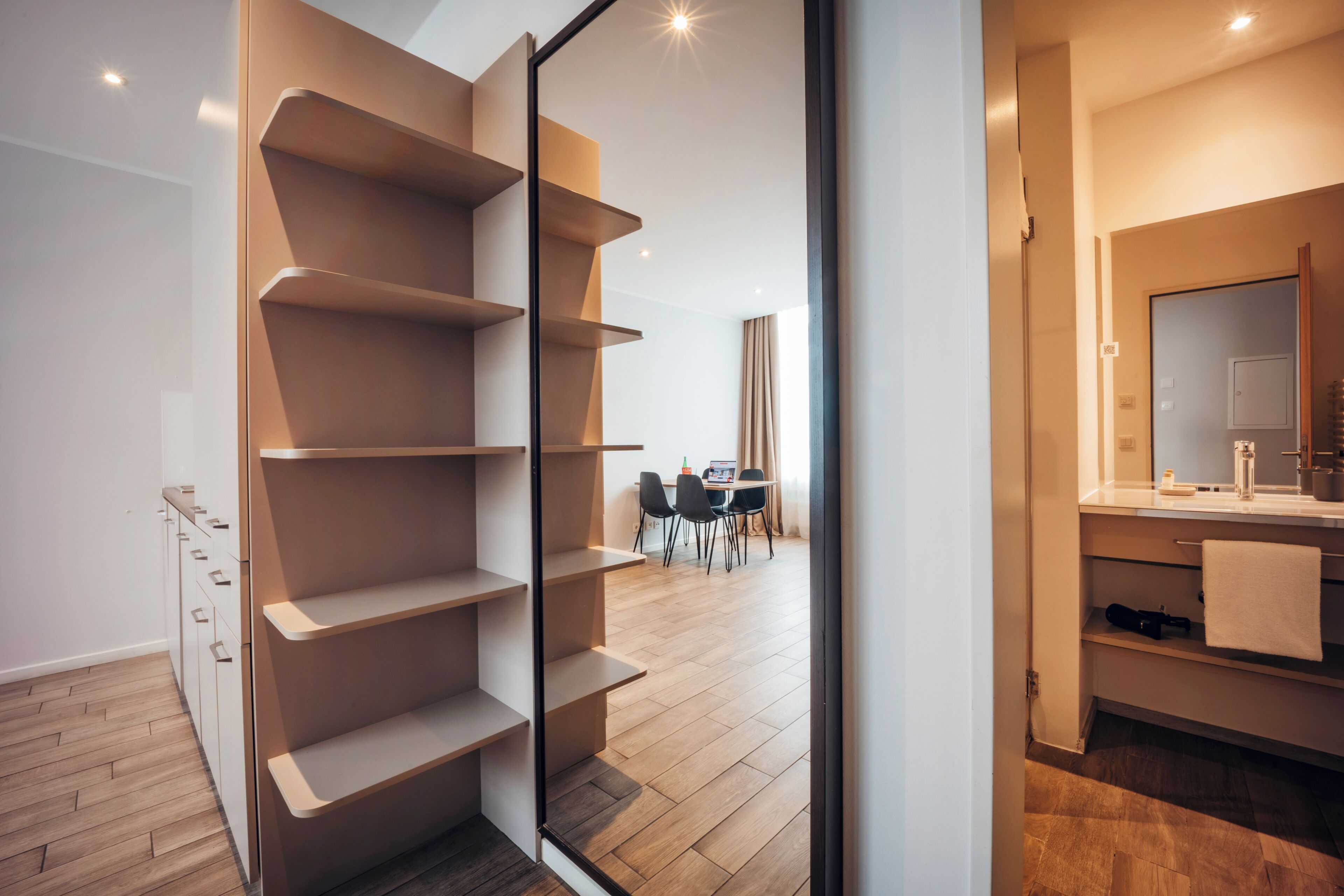 A modern interior with a built-in shelving unit, a dining area with chairs, and a bathroom with a mirror and sink.