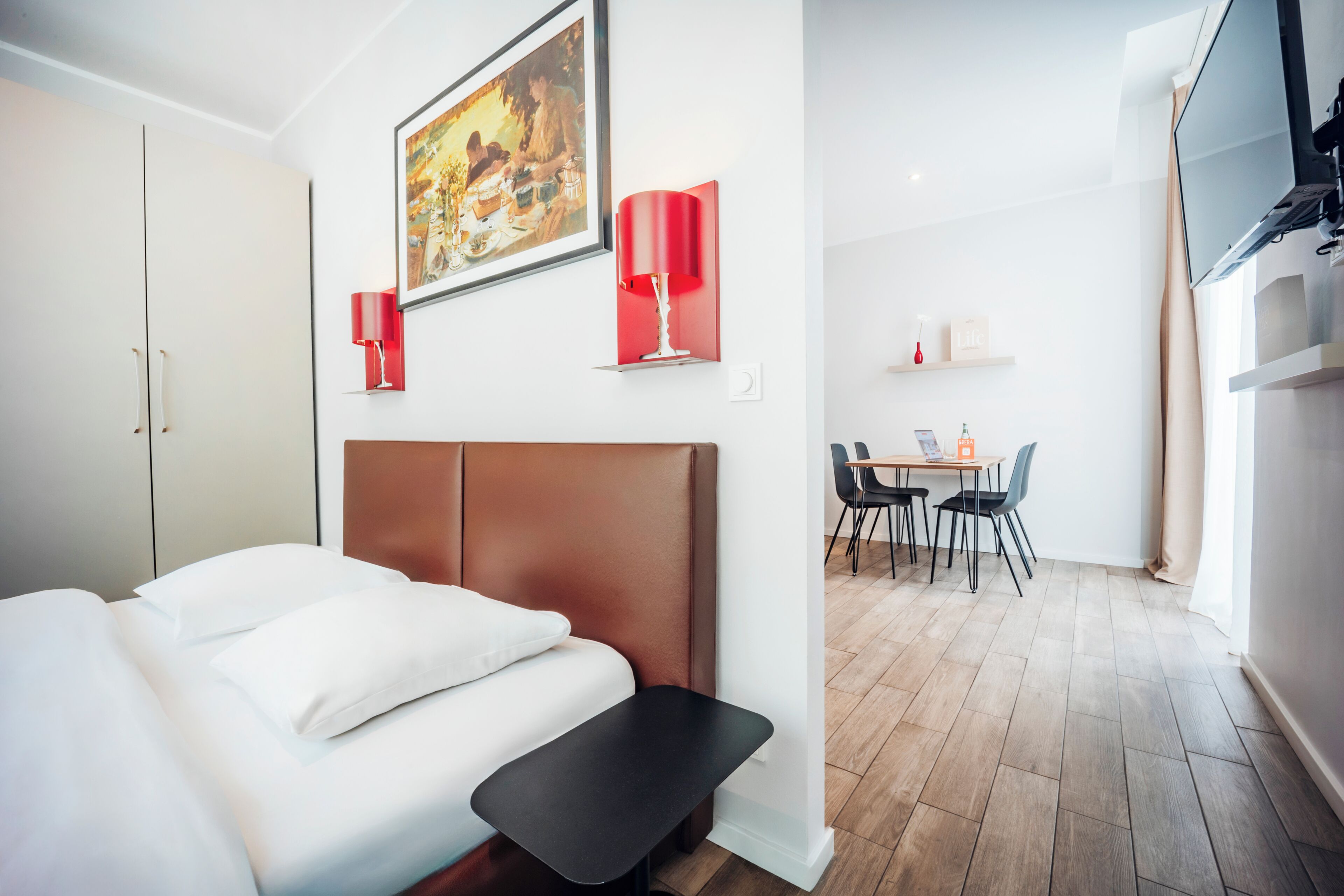 A modern hotel room with a bed, a dining area with chairs and a table, and a kitchen area with a stove.