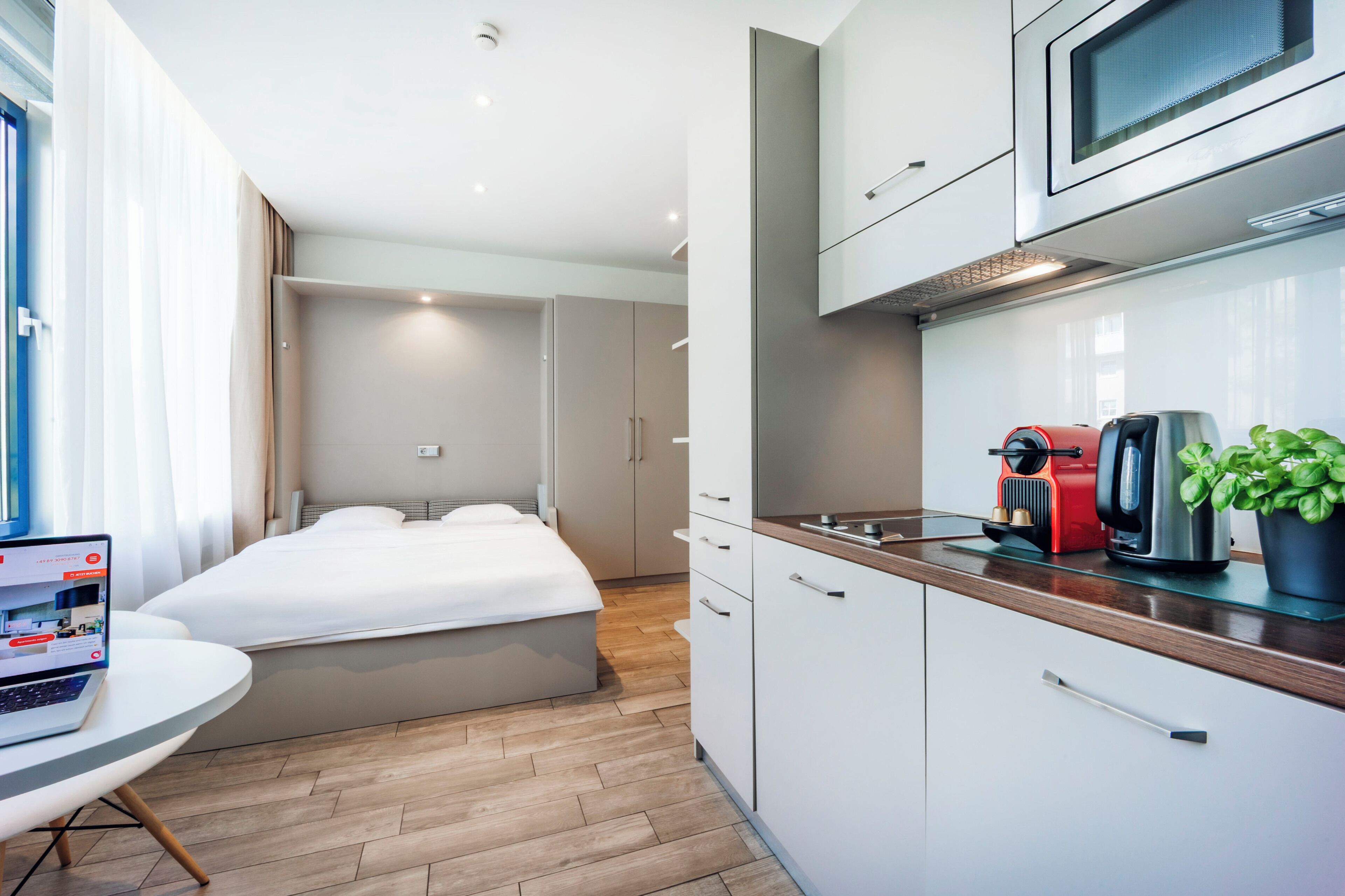 Moderni hotellihuone, jossa on sänky, työpöytä kannettavalla tietokoneella, pieni keittiönurkkaus mikroaaltouunilla ja ruukkukasvi.