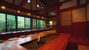 Una habitación tradicional japonesa con suelo de tatami, una baja mesa de madera con cojines y vistas a un bosque a través de amplios ventanales.