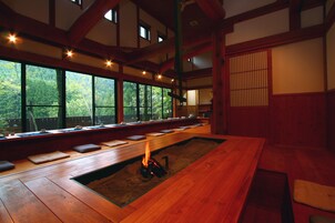 Una habitación tradicional japonesa con suelo de tatami, una baja mesa de madera con cojines y vistas a un bosque a través de amplios ventanales.