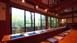 Un largo banco de madera con cojines, una mesa de madera con tazas y platillos, y grandes ventanales que ofrecen vistas a un área boscosa.