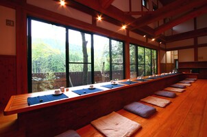 Un largo banco de madera con cojines, una mesa de madera con tazas y platillos, y grandes ventanales que ofrecen vistas a un área boscosa.
