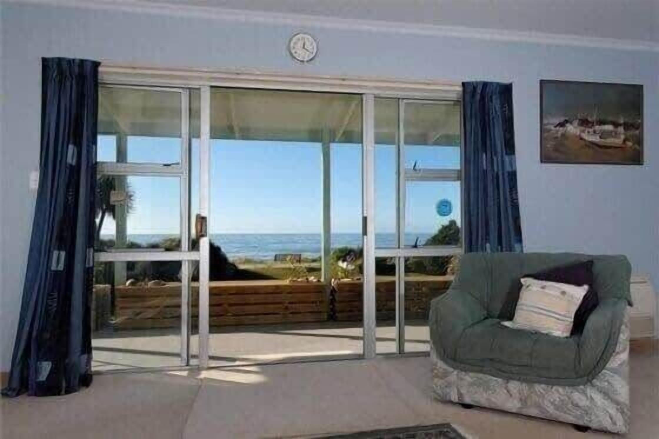 A room with a large sliding glass door leading to a balcony with a view of the sea. There is a green armchair with a cushion and a clock on the wall.