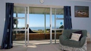 A room with a large sliding glass door leading to a balcony with a view of the sea. There is a green armchair with a cushion and a clock on the wall. - Sunrise at Parapara Beach (Parapara)