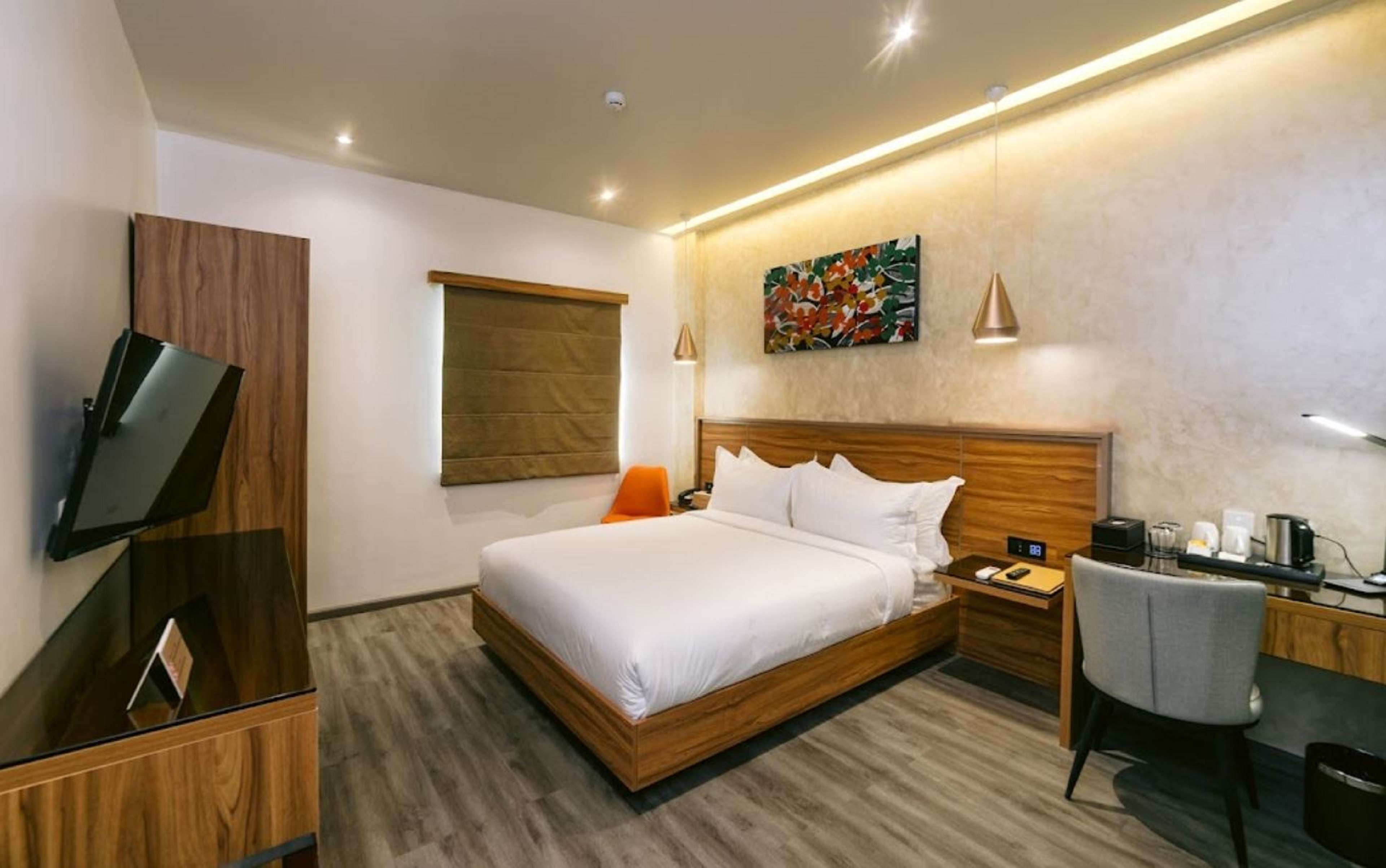 A modern hotel room with a wooden bed, a desk with a chair, a TV, and a painting on the wall.
