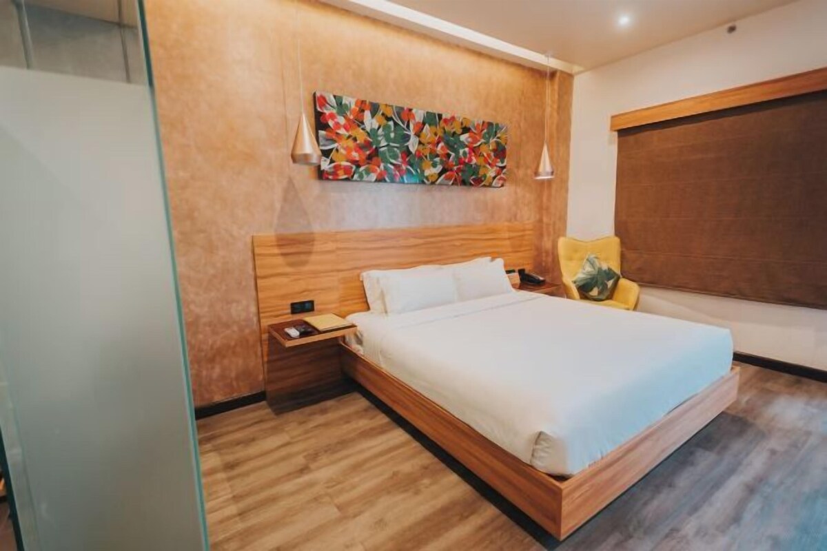 A modern hotel room with a wooden headboard, a large bed, a nightstand, a bedside table, a colorful abstract painting on the wall, and a large window with a view.