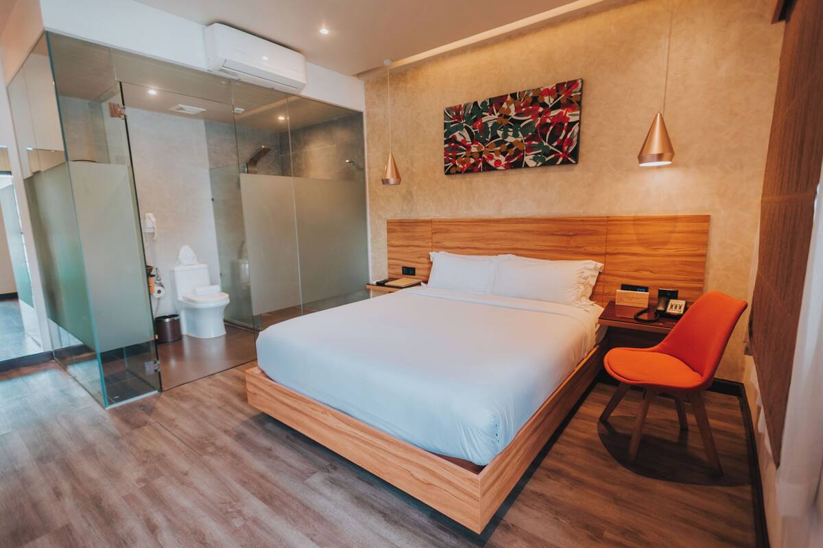 A modern hotel room with a wooden bed, a glass shower enclosure, a wooden nightstand, and a red chair.