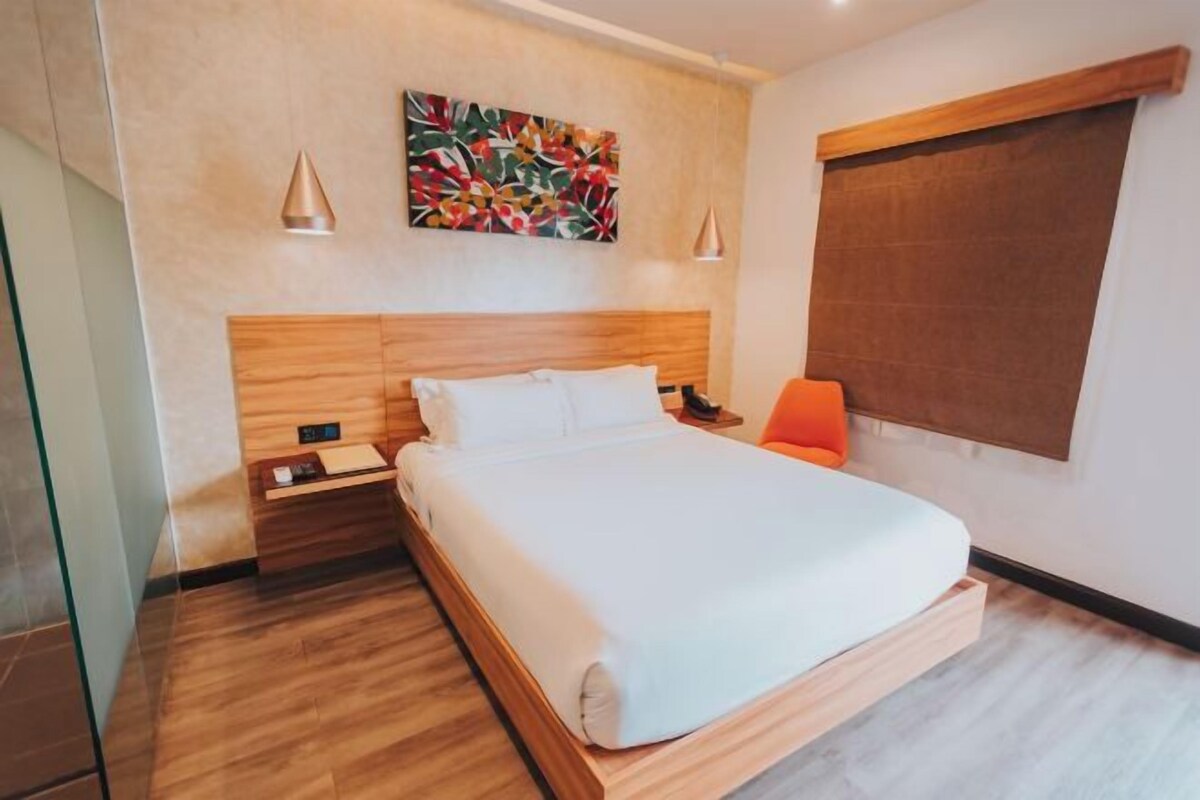A modern hotel room with a wooden bed, a nightstand, a bedside table, a colorful abstract painting, and a window with a brown blind.