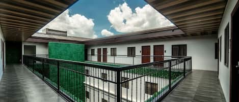 Un espacio al aire libre cubierto con vista al cielo a través de una abertura triangular, rodeado por un edificio blanco con múltiples puertas y ventanas, y una pared verde.