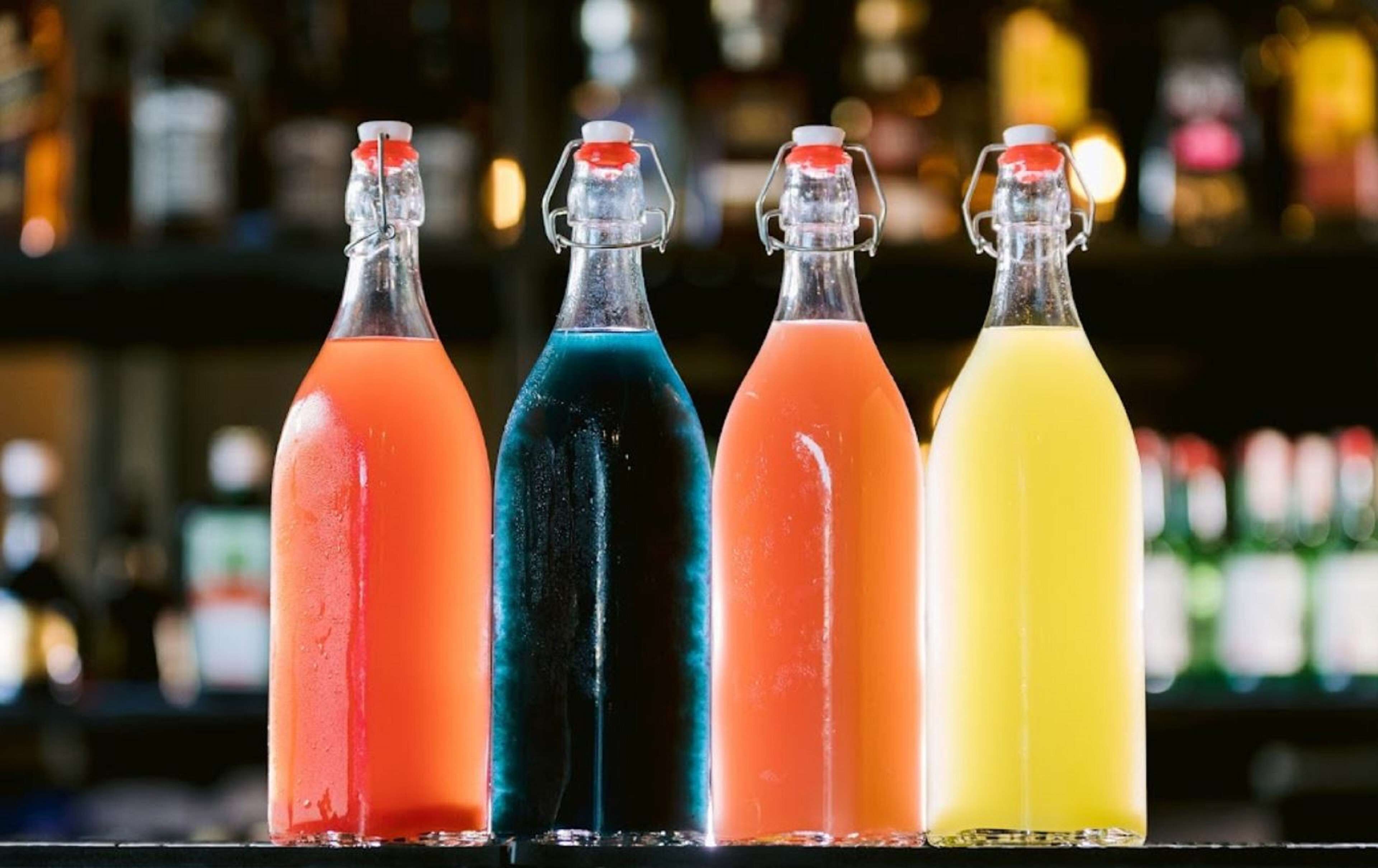 Cuatro botellas de bebidas de colores sobre una barra, con un fondo desenfocado de otras botellas.