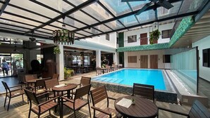 Uma piscina interior com teto de vidro, rodeada por mesas e cadeiras de madeira.