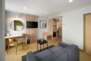 A modern hotel room with a wooden desk, a grey sofa, a flat-screen TV, and a mirror.
