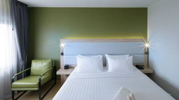 Quarto de hotel com cama, uma cadeira verde, duas mesas de cabeceira com luminárias e uma janela com cortinas.