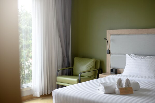 A hotel room with a bed, a green chair, a window with curtains, and a bedside table with a lamp.