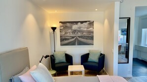 Un dormitorio moderno con una cama grande, dos sillones, una mesita y un cuadro de un camino en la pared.