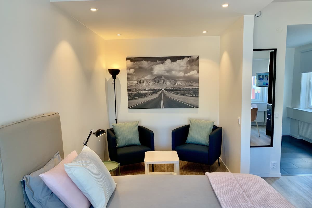 Un dormitorio moderno con una cama grande, dos sillones, una mesita y un cuadro de un camino en la pared.