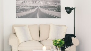 Un salón con un sofá color beige, una lámpara de pie negra, un retrato enmarcado de una carretera, y una mesita con una vela y flores.