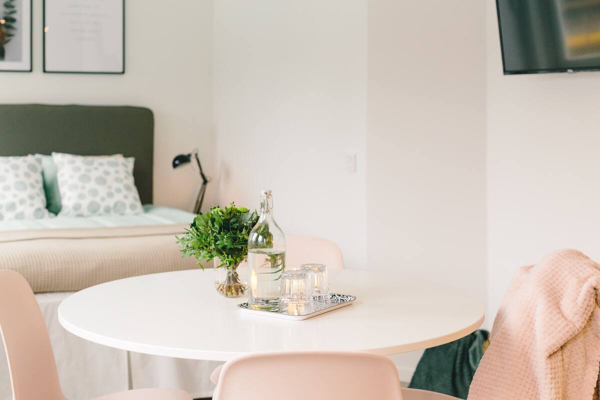 Una habitación ordenada con una mesa redonda blanca, una cama con almohadas verdes y blancas, una planta pequeña y una botella.