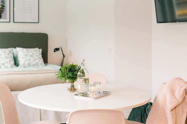 Una habitación ordenada con una mesa redonda blanca, una cama con almohadas verdes y blancas, una planta pequeña y una botella.