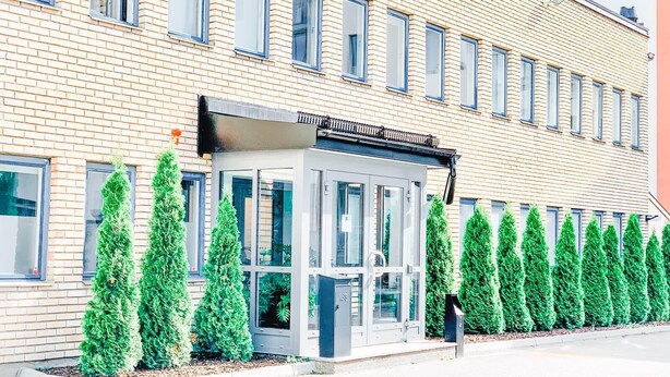 Un edificio moderno con fachada de ladrillo, entrada de vidrio y una fila de árboles verdes al frente.