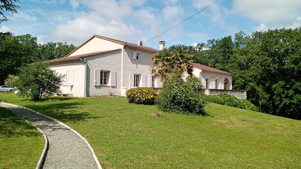 Une maison à deux étages, avec un toit recouvert de tuiles, entourée d’une végétation luxuriante et d’une pelouse bien entretenue.