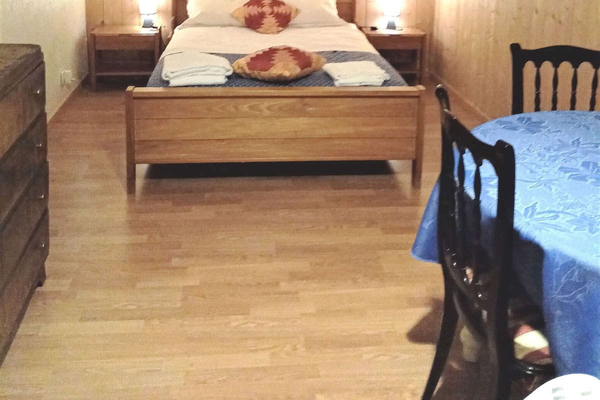 A small, cozy room with a wooden bed, two bedside tables, a chair, and a table with a blue tablecloth.
