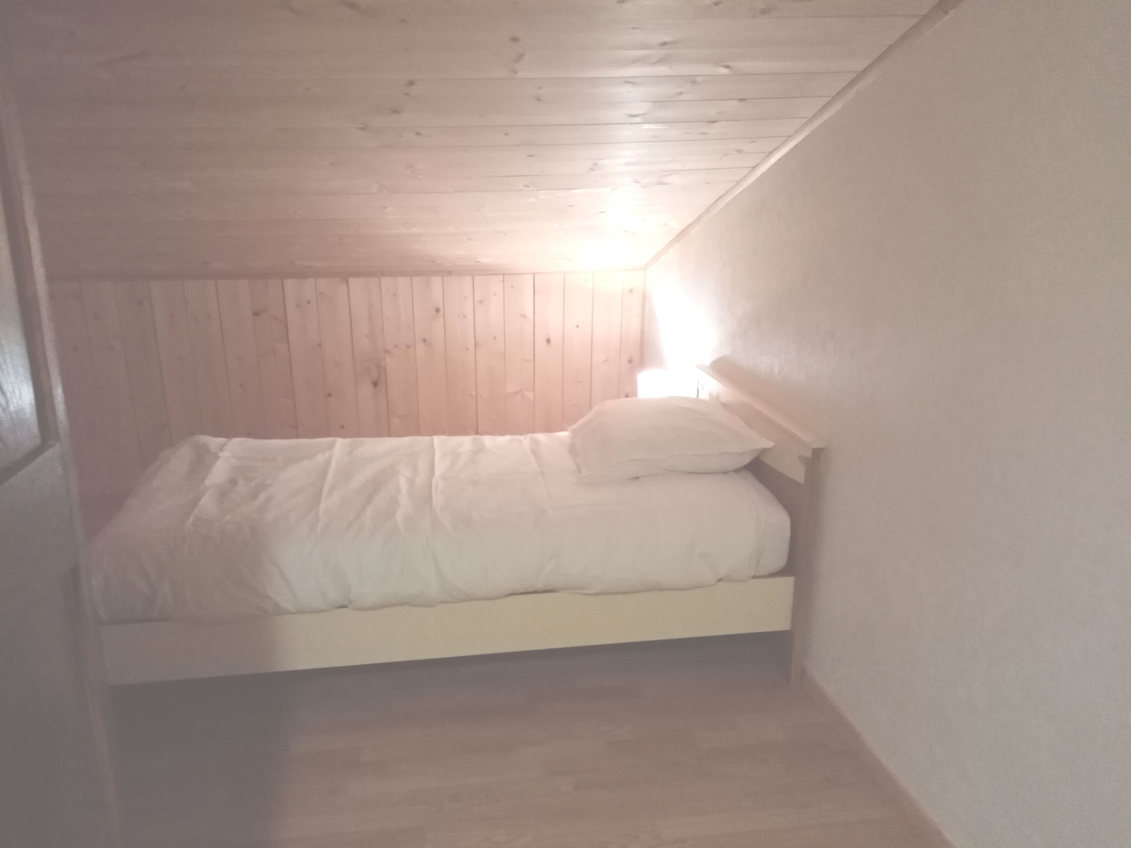 A small, dimly lit attic bedroom with a wooden ceiling and walls, a single bed with white bedding, and a small bedside table.