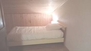 A small, dimly lit attic bedroom with a wooden ceiling and walls, a single bed with white bedding, and a small bedside table.