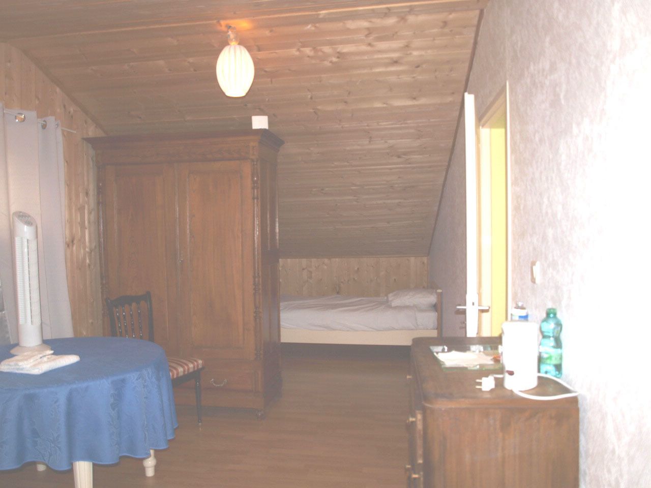 Une petite pièce faiblement éclairée, avec un lit simple, une table recouverte d’une nappe bleue et une commode en bois.