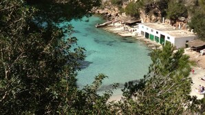 Zona costera con un mar turquesa cristalino, terreno rocoso y un edificio con puertas verdes.