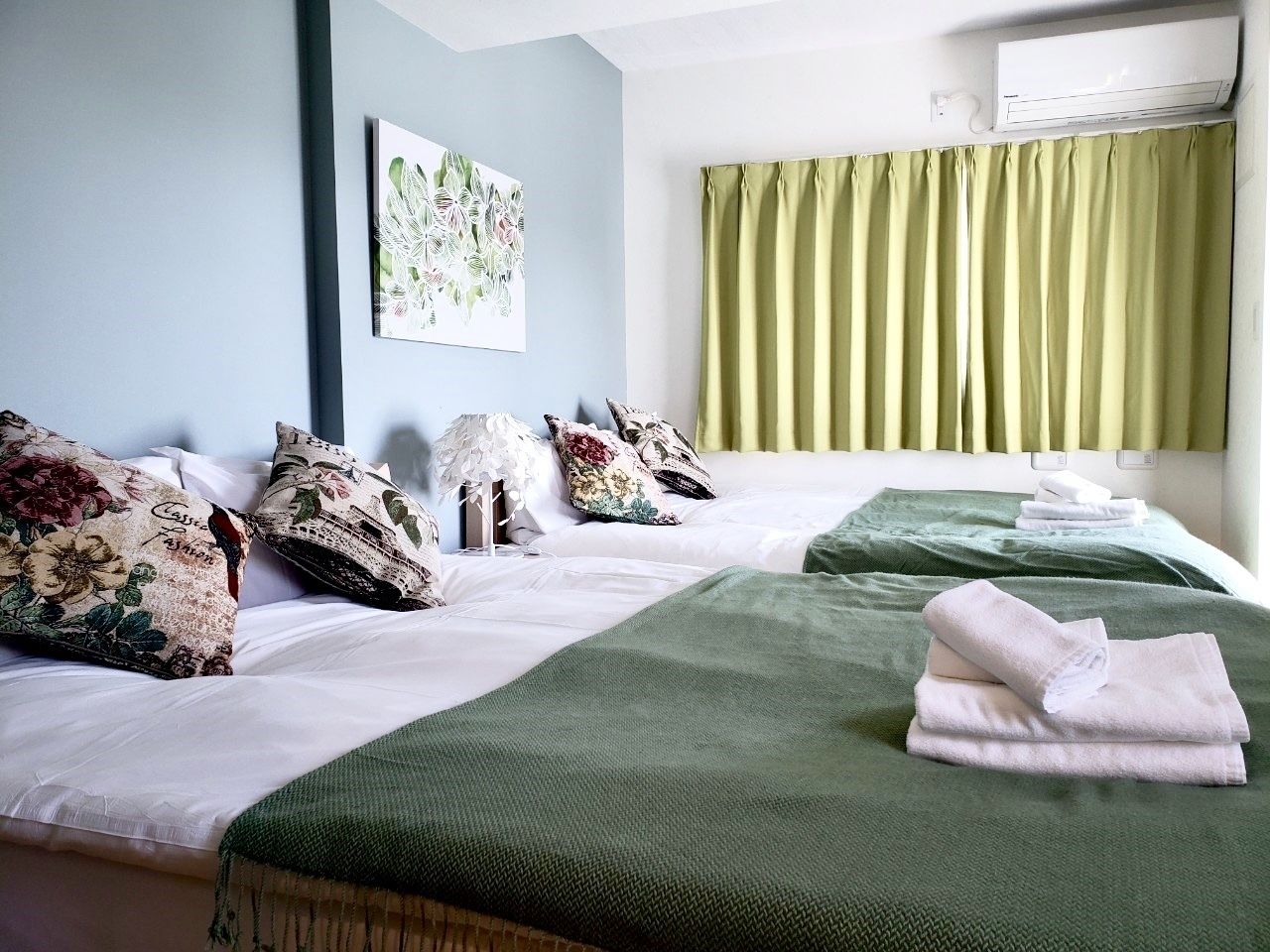 Une chambre d’hôtel avec deux lits, des oreillers à motifs floraux, une literie verte, un climatiseur fixé au mur et une fenêtre avec des rideaux verts.
