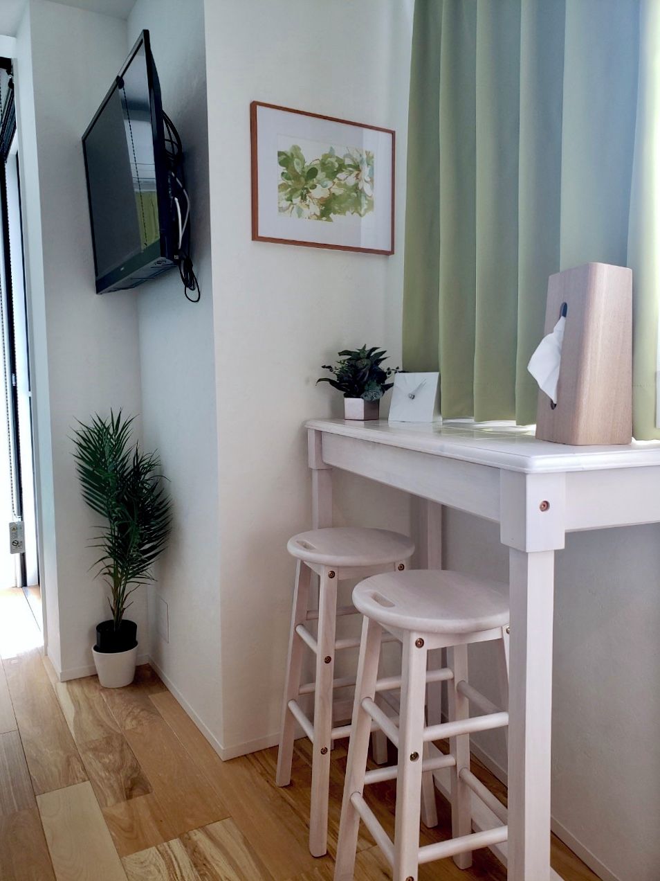 A small, white desk with two stools, a framed picture on the wall, and a potted plant beside it.