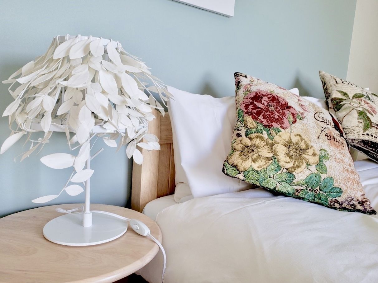 Une chambre à coucher bien rangée, avec une table de chevet ronde en bois, une lampe blanche décorée de motifs de feuilles et un lit agrémenté d’oreillers à motifs floraux.