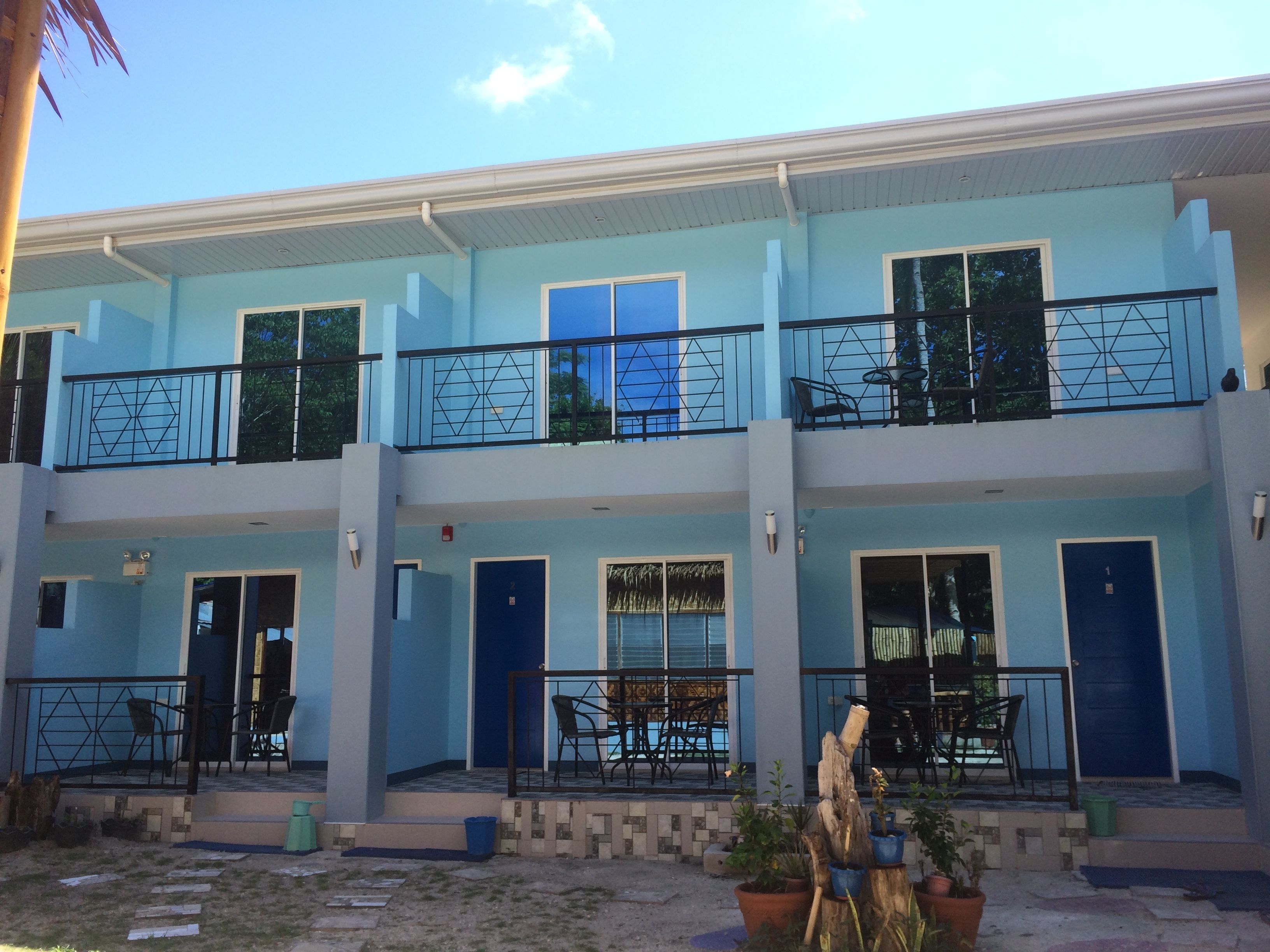 Un edificio de dos pisos con fachada azul claro, múltiples balcones con barandillas negras y varias puertas y ventanas.