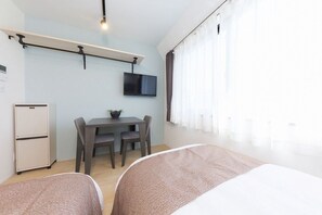 Habitación de hotel pequeña con cama, escritorio con dos sillas, televisor montado en la pared y refrigerador.