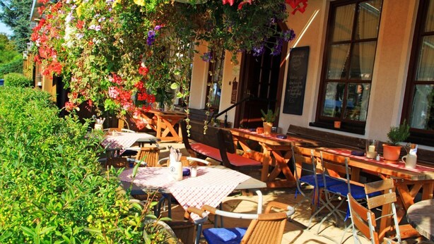An outdoor café with tables and chairs, surrounded by greenery and a building with large windows. - Hotel Häußler (Reichertshofen)