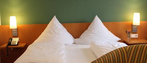 Una habitación de hotel con una cama bien hecha, dos lámparas de noche, un teléfono, una mesita con un florero y un cuadro en la pared.