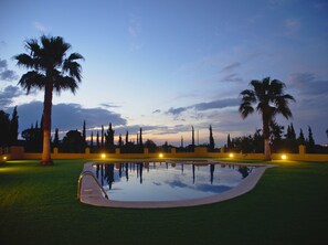 Piscina com um calçadão ao redor, palmeiras e céu ao pôr do sol.