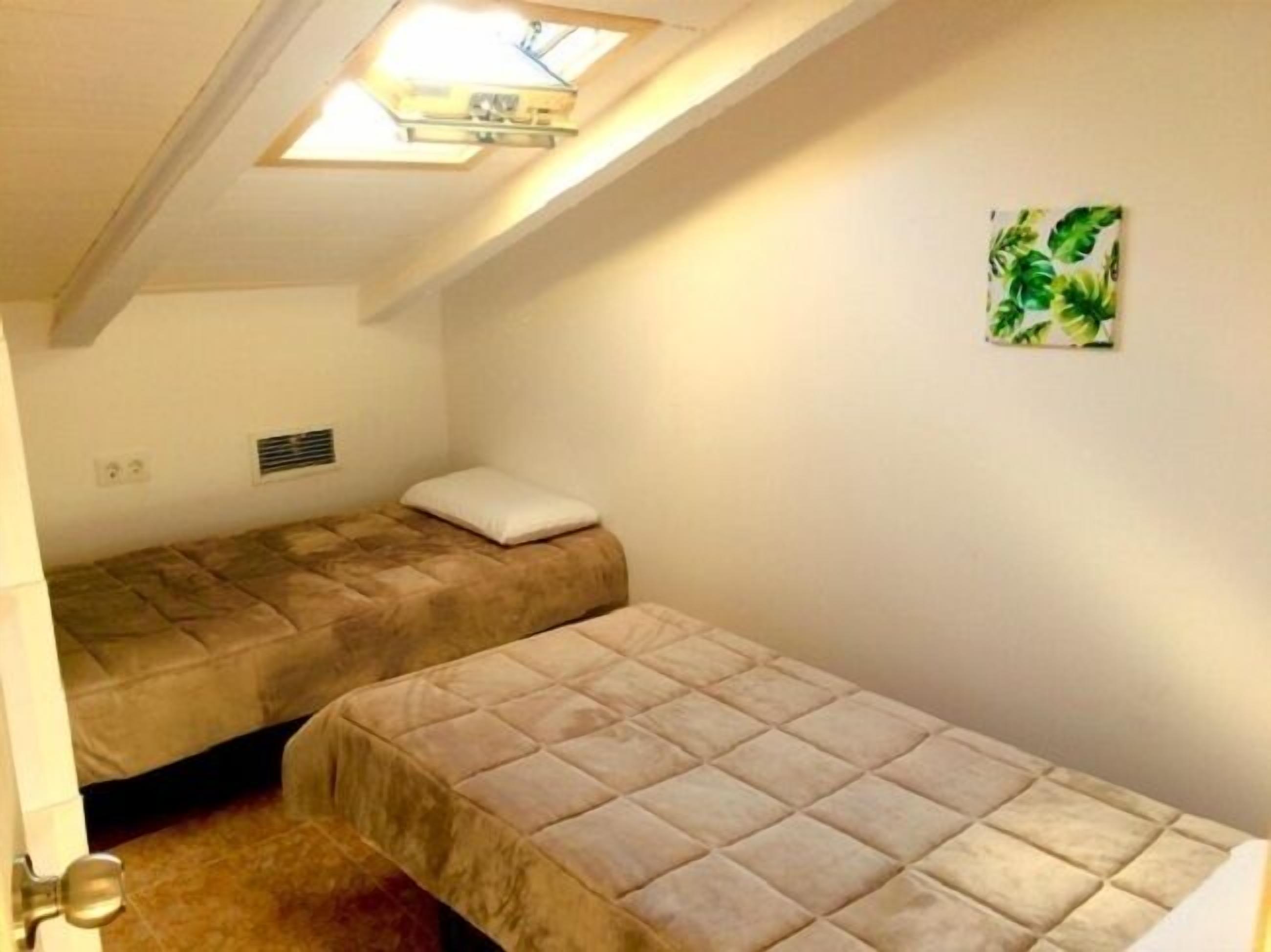 Una habitación pequeña con dos camas, un tragaluz, un respiradero en la pared y un cuadro con temática tropical.