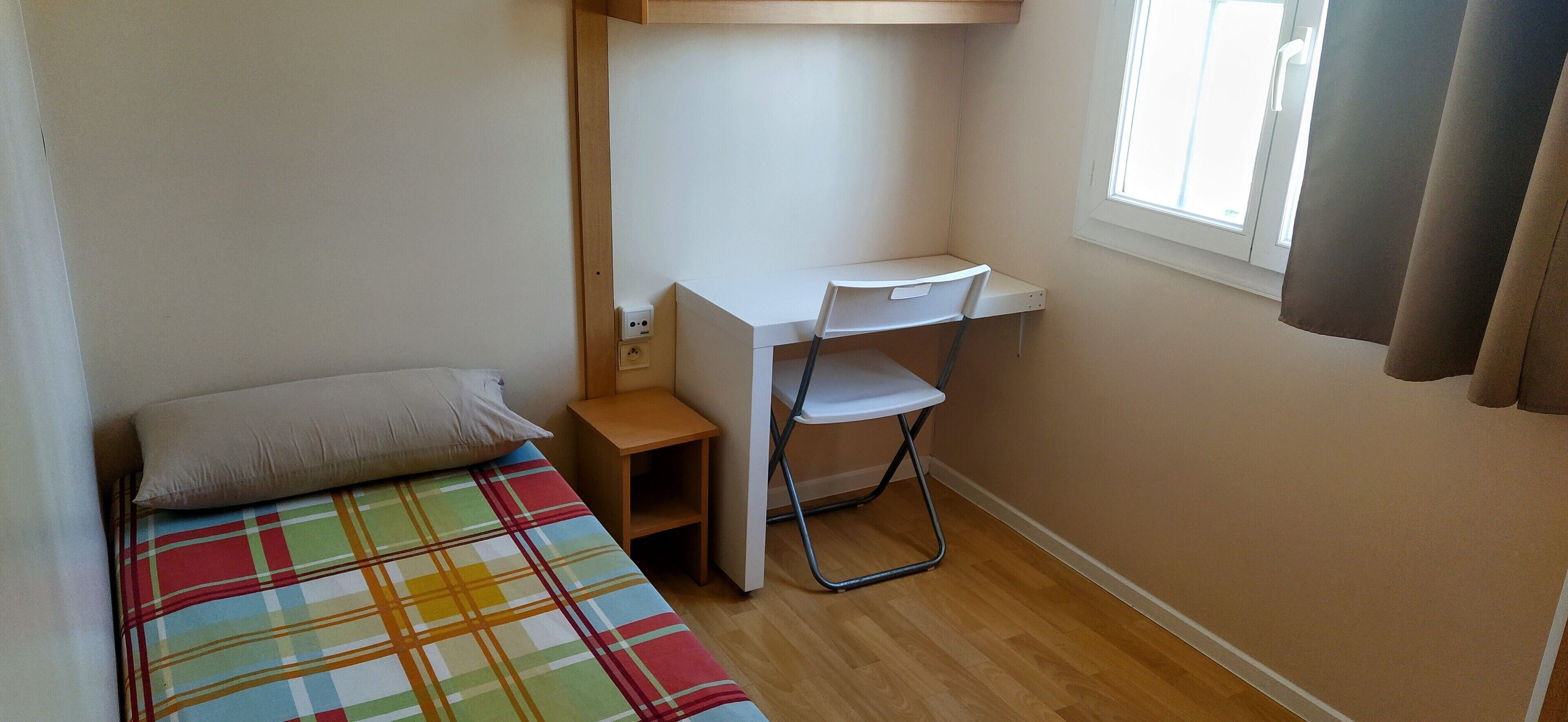 Una habitación pequeña con cama individual, colcha colorida, escritorio con silla y ventana con cortinas.