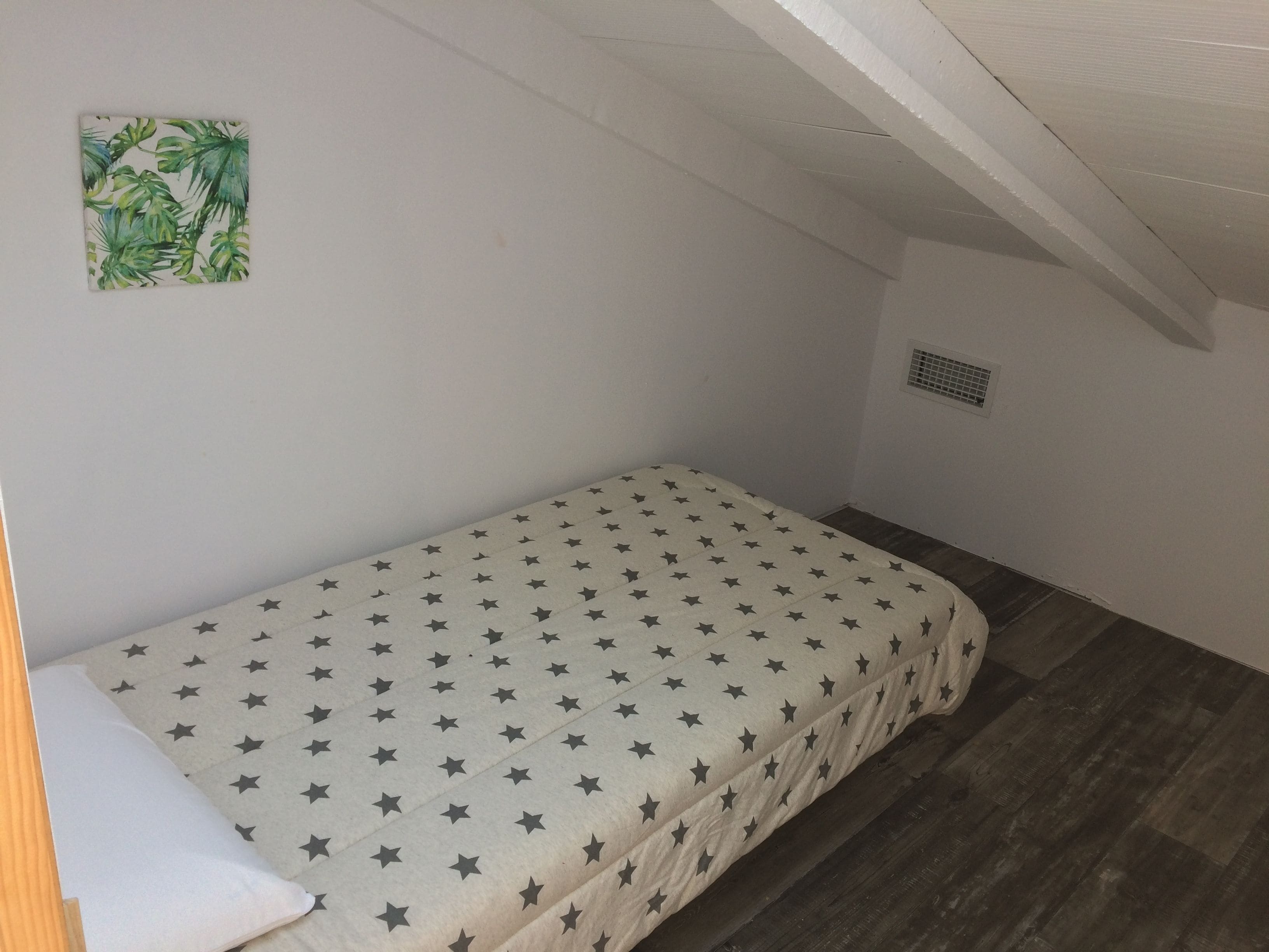 Quarto pequeno com uma cama de solteiro, colcha com padrão de estrelas, travesseiro branco e uma pequena planta na parede.